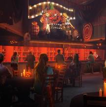 an indoors atmospheric bar setting, with clown characters overhead on a platform performing and patrons observing from below.